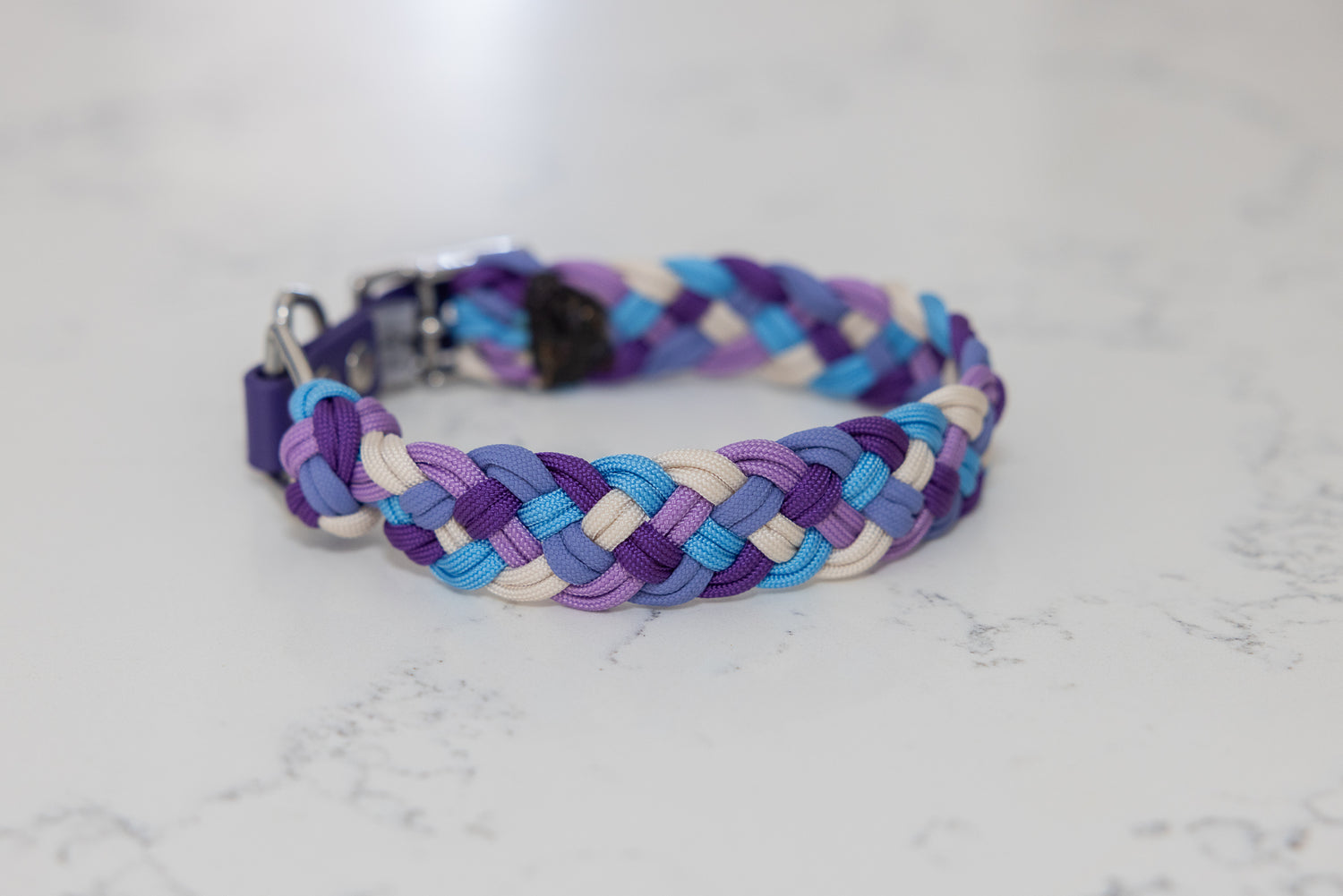 A multicolored paracord dog collar with purples, blue and cream strand displayed on a marble background.