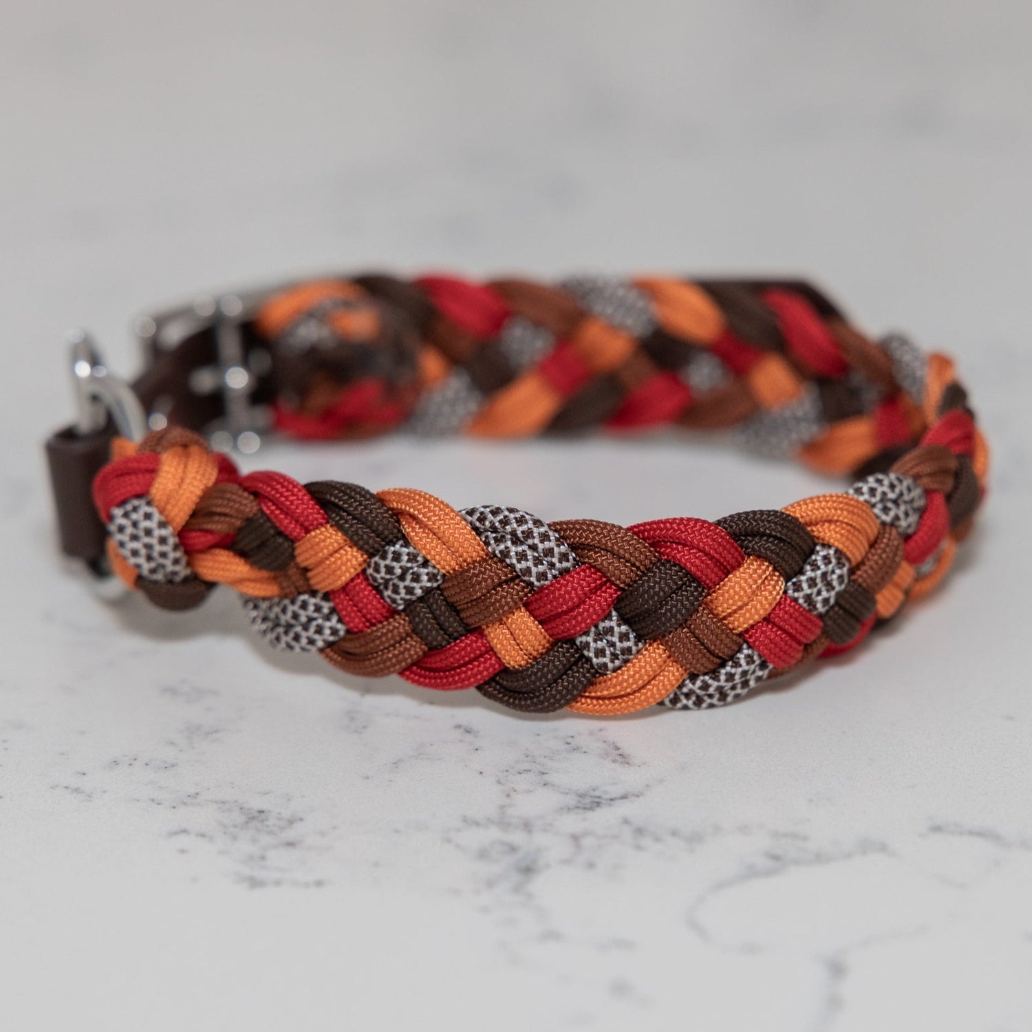 A braided paracord collar with a metal buckle, featuring a pattern of red, orange, and brown colors.